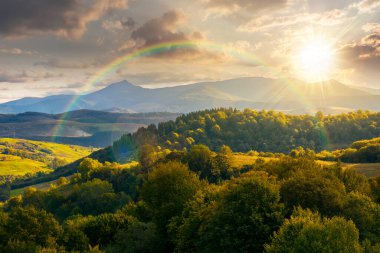Eylül günbatımında kırsal alan. Vadideki köy ve ormanlık tepelerdeki kırsal araziler akşam ışığı altında. Gökkuşağının altında yüksek zirvesi olan dağ sırtı