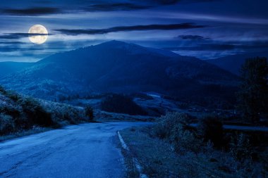 Gece dağ geçidi. Asfalt yol tepeden aşağı sarkıyor. Dolunay ışığında bulutlu gökyüzünün altında dağın önündeki vadiyle kırsal bir manzara.