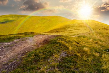 Yaz aylarında gün batımında dağ turizmine giden yol. Akşam ışığında alp manzarası. Gökkuşağının altında yeşil tepeler ve bulutlu mavi gökyüzü olan güzel bir tepe manzarası.