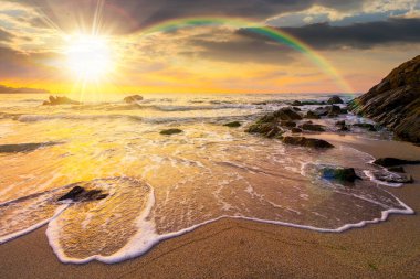 Gün batımında körfezdeki kayaların üzerinde gökkuşağı olan deniz kıyısı. Deniz gelgiti ve sakin dalgalar akşam ışığında altın kumlu sahili yıkıyor. Balayı için deniz kenarında güzel bir sonbahar tatili.