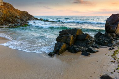 Yazın gün doğumunda deniz kenarındaki manzara. Turuncu ışıkta sakin dalgalarla yıkanan kayalarla dolu güzel bir sahil. Mavi gökyüzünün altında bulutlarla birlikte ufuk çizgisine bak. Rahatlamak ve rahatlamak için manzaralı seyahat yeri