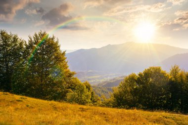 Sonbaharda günbatımında tepenin üzerinde ormanla kaplı dağ manzarası. Akşam ışığında kırsal vadiye güzel bir manzara. Gökkuşağı ve gökyüzü altında bulutlu dağlık arazi manzarası