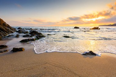 Sahilde gün doğumu. Deniz tatili için güzel bir yaz manzarası. Kumda kayalar. Sudaki sakin dalgalar. Gökyüzündeki bulutların arkasında güneş. Sozopol 'daki bambu plajının geniş panoramik manzarası