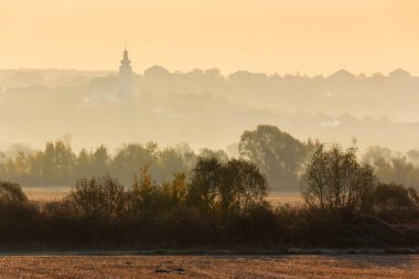Kırsal alan bir sonbahar sabahında. Boş tarlalar boyunca sıra sıra ağaçlar. Tepede kilisesi olan bir köy ormanın arkasında sisli bir mesafede.