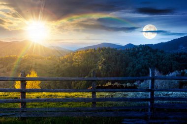 trees on the hill and fence on the grassy field. day and night time change concept. spring rural landscape. distant mountain ridge beneath sky with sun and moon at twilight. idyllic alpine farmland