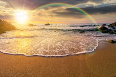 Gün batımında körfezdeki kayaların üzerinde gökkuşağı olan deniz kıyısı. Deniz gelgiti ve sakin dalgalar akşam ışığında altın kumlu sahili yıkıyor. Balayı için deniz kenarında güzel bir sonbahar tatili.