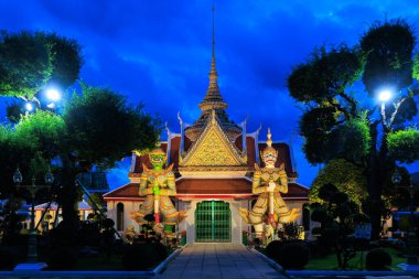 Wat Arun Tapınağı ana girişi önünde iki şeytan heykeli var. Bina gece aydınlanıyor. Bangkok