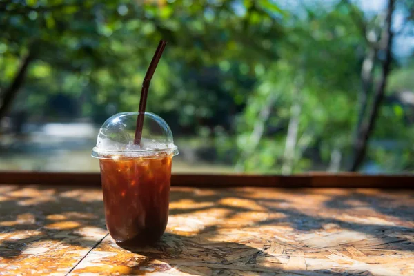 Ice Americano coffee in plastic cup on wooden table with blur natural river and tree. Cafes in greenery environment.
