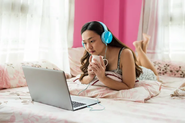 Beautiful Asian young woman on bed watch streaming online movie, music video from laptop computer and drink hot coffee in morning. leisure lifestyle at home bedroom.