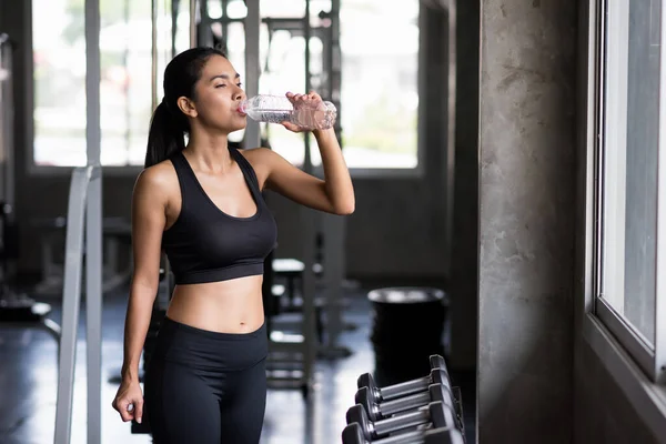 Attractive Asian fit young woman break and drink water after dumbbell muscle exercise in fitness gym. Refreshment after muscular bodybuilding and hHealthy lifestyle concept.