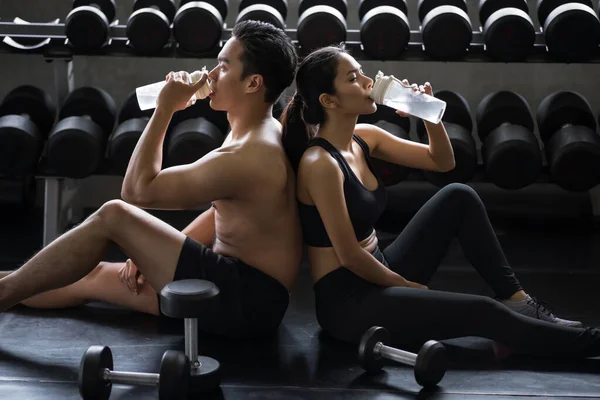 Athletic young fit Asian couple with sexy muscle body break and drink water after dumbbell work out or exercise in fitness gym. Bodybuilding and healthy lifestyle concept.