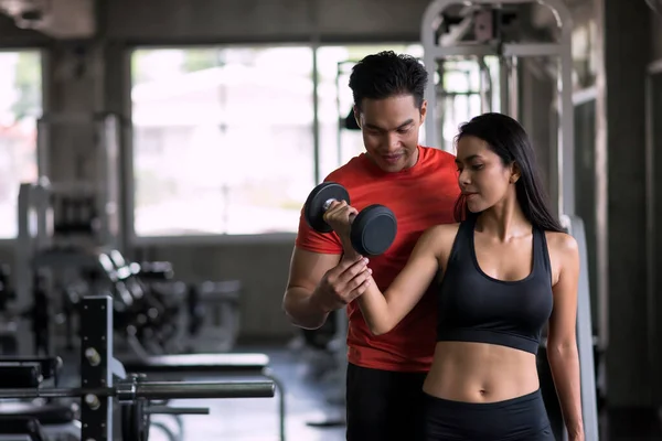 Portrait of personal trainer or boyfriend coach dumbbell exercise to sexy woman in sport fitness gym. Happy couple working out for bodybuilding and healthy lifestyle.