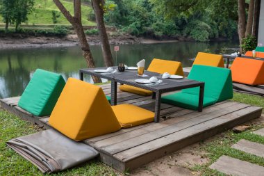 Outdoor restaurant table and colorful sofa seats by kwai river with peaceful atmosphere in Kanchanaburi, Thailand.