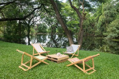 Wooden deckchair and table on green grass garden by pond with beautiful big trees and reflection on water. Outdoor cafe and restaurant.