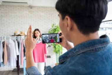 Behind the scene concept. Record live streaming by mobile or smartphone to sell dress on social media by beauty blogger. Videographer give hand sign to female influencer.
