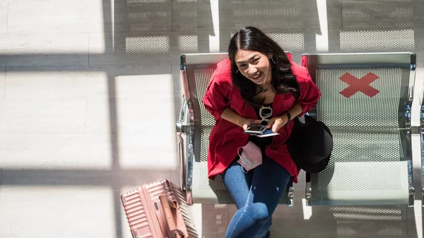 Top view of beautiful smiling Asian tourist woman sit on waiting seat with luggage. Wait for departure in airport terminal. New normal to prevent covid-19 omicron pandemic.