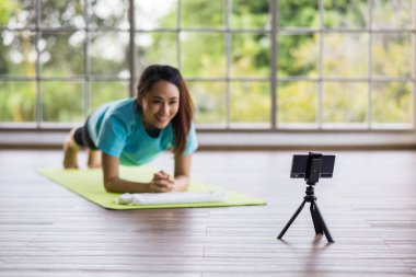 Genç Asya yogası gülen antrenörü cep telefonu ve tripodla kaydedilen çevrimiçi egzersiz kursunu gösteriyor. Akıllı telefonlara bakıyor. Telefona odaklan..