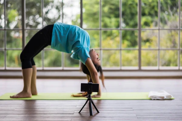 Blur sport influencer blogger record practicing yoga to live streaming online. Asian woman standing in Urdhva Dhanurasana exercise, Bridge pose. Focus at smartphone.