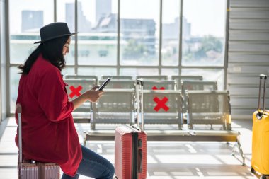 Güzel, genç Asyalı bir kadın havaalanının kalkış terminalinde bagaja oturup akıllı telefon ya da mobie telefon çalar. Covid-19 omikron salgınını önlemek için yeni normallerle seyahat et.