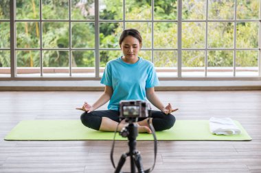 Genç Asyalı kadın blogcu Lotus avuç içi meditasyon yogası yapıyor ve gözlerini kapatıyor. Çevrimiçi sosyal medya kanalından dijital kamerayla spor etkinleştirici canlı ders.