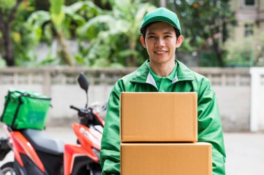 Yeşil üniformalı Asyalı gülen sürücü eve motosikletle paket teslim ediyor. Müşteriye göndermek için el ele tutuşma kutusu.