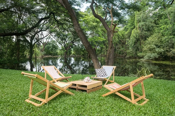 Relax wooden deckchair and table on green grass yard by pond with beautiful big trees and reflection on water. Outdoor cafe and restaurant.