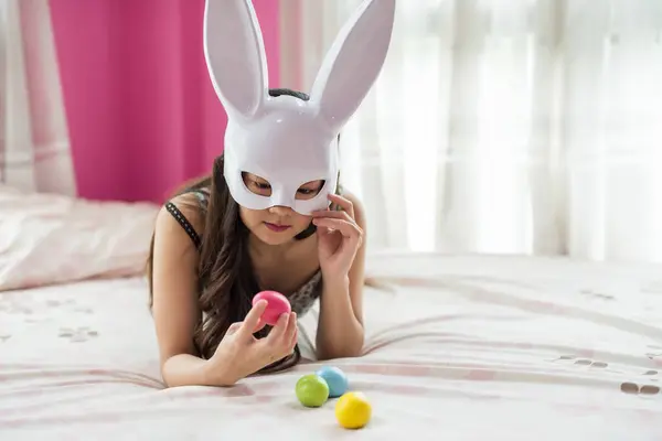 Young Asian woman in white bunny mask on bed grab and look at pink eggs on hands. Portrait of Chinese girl celebrate Easter holiday 2024 in bedroom.