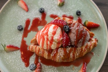 Yakın plan vanilyalı ve çikolatalı kruvasan üzerine çilek soslu dondurma ve ahşap masada yabanmersini. Tatlı mı, sağlıksız mı?.