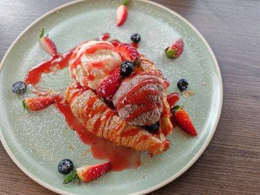 Vanilya ve çikolatalı kruvasan üzerinde çilek soslu dondurma ve ahşap masada yabanmersini. Tatlı mı, sağlıksız mı?.