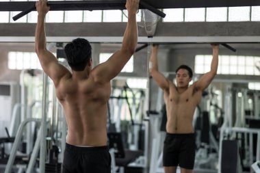 Spor salonunda kaslı atlet Asyalı adam aynanın önünde yatay çubuğa yanaşıyor. Üstsüz adamın sırt kasları sıkışıyor ve kendine bakıyor. Spor vücut geliştirme.