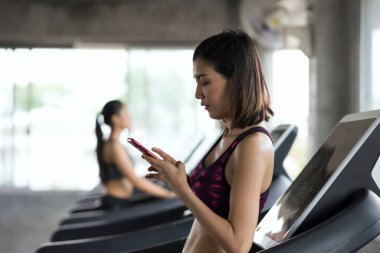 Güzel formda bir Asyalı genç kadın koşu bandında koşmaktan ve spor salonunda cep telefonu kullanmaktan kaçıyor. Vücut geliştirme ve spor sağlıklı yaşam tarzı konsepti.