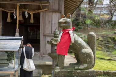 Kitsune fox statue with woman pray at Homangu Kamado shrine, Dazaifu, Kyushu., Japan. Holy trickster foxes from Japanese folklore. Shrine inspire for Kimetsu no Yaiba: Demon Slayer. clipart