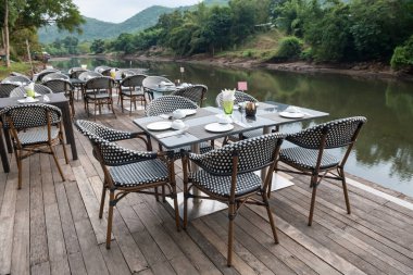 Kanchanaburi, Tayland 'da huzurlu bir atmosferi olan Kwai nehrinin kıyısındaki ahşap teras üzerindeki açık hava restoran masası ve benekli sandalyeler..