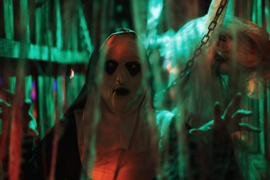 Kafesteki korkunç şeytani rahibe. Gelini asmış, gelinlik giymiş, yeşil ışık yanıyor. 2025 Cadılar Bayramı konsepti parti maskesi ve kostüm .