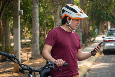 Beyaz kasklı, kırmızı tişörtlü, kahverengi pantolonlu, elektrikli bisiklete binen ve telefonuna bakan Latin bir adam.