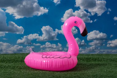 Pembe, şişirilebilir bir flamingo havuzu berrak mavi bir gökyüzünün, kabarık beyaz bulutlarla kaplı bir zeminine karşı canlı bir şekilde duruyor.