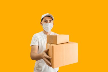 Asyalı genç teslimatçı şapkalı ve beyaz tişörtlü sarı arka planda bir paket posta kutusu tutuyor. hızlı dağıtım hizmeti kavramı.