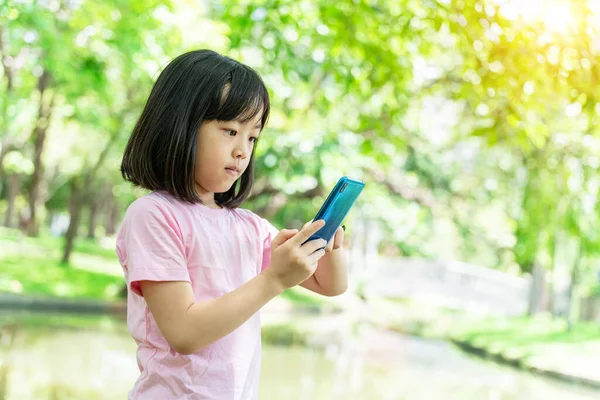 stock image Beautiful asian child girl using mobile smartphone. Little girl is playing in digital game in the green park.