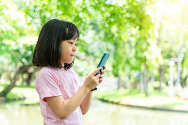 Güzel Asyalı kız çocuğu cep telefonu kullanıyor. Küçük kız yeşil parktaki dijital oyunda oynuyor..