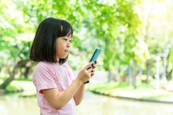 Güzel Asyalı kız çocuğu cep telefonu kullanıyor. Küçük kız yeşil parktaki dijital oyunda oynuyor..