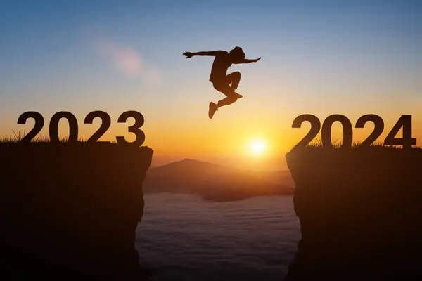新年2024コンセプト 人のシルエットは日没または日の出の背景の上に2023年から2024年の間崖にジャンプします ロイヤリティフリーのストック画像