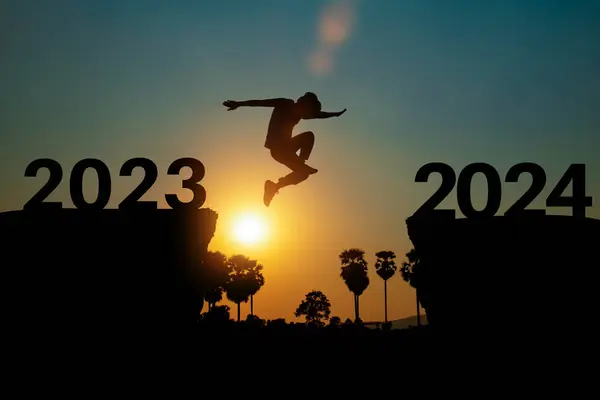 新年2024コンセプト 人のシルエットは日没または日の出の背景の上に2023年から2024年の間崖にジャンプします ロイヤリティフリーのストック写真
