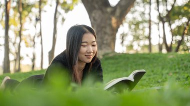 Güzel Asyalı kız çimenlerde uzanıyor ve parkta kitap okuyor, rahatlama kavramı..