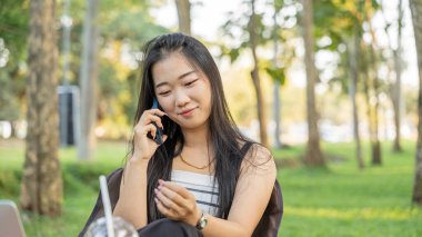 Güzel gülümseyen Asyalı kız parkta akıllı telefonla konuşuyor.