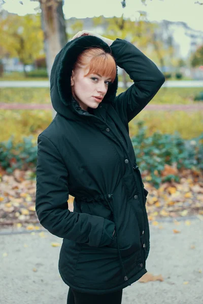 Redhead woman in hooded black coat posing at autumn during forest walk
