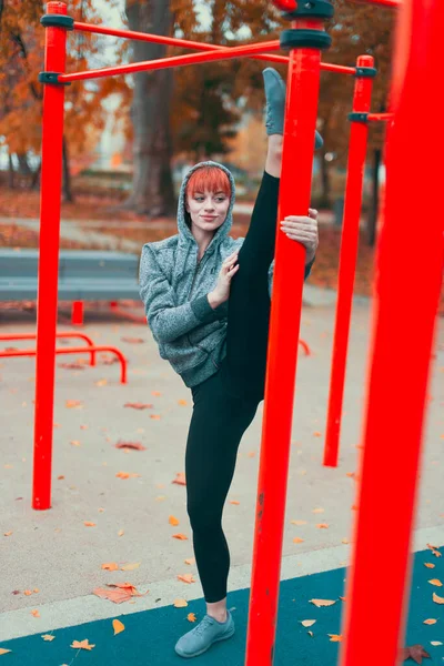 Young hooded Caucasian ballerina woman warming up at playground pillars and doing splits at autumn in park
