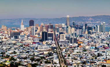 San Francisco şehir merkezindeki gökdelenler ve Market Sokağı koridoru boyunca uzanan tarihi binalarla panoramik manzara aydınlık bir yaz günü arka planda Doğu Körfezi tepeleri ile yukarıdan görülüyor.
