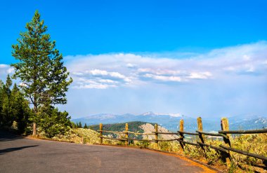 Grand Teton Ulusal Parkı 'ndaki Sinyal Dağı yakınlarında panoramik bir bakış açısı, yolun kenarını çevreleyen orman tepeleri ve uzak tepeleri canlı bir yaz gökyüzü altında gösteren ahşap bir çiti gösteriyor..