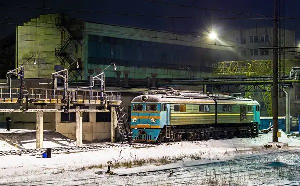 Ukrayna 'daki Lviv Lokomotif Tamir Tesisi' nde karlı raylara park edilmiş eski bir Sovyet lokomotifinin gece görüşü. Soğuk sanayi sahnesinde beton yapılar, soluk turkuaz ve turuncu çizgili yıpranmış bir lokomotif ve dramatik