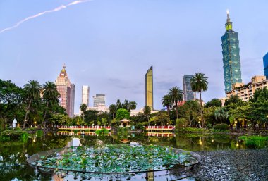 Tayvan 'daki modern gökdelenlerin şehrin üzerinde yükseldiği akşam manzarası, Nilüfer bitkileri ve palmiye ağaçlarıyla çevrili Sun Yat Sen Anıt Parkı' nın sakin göletine yansıyor.
